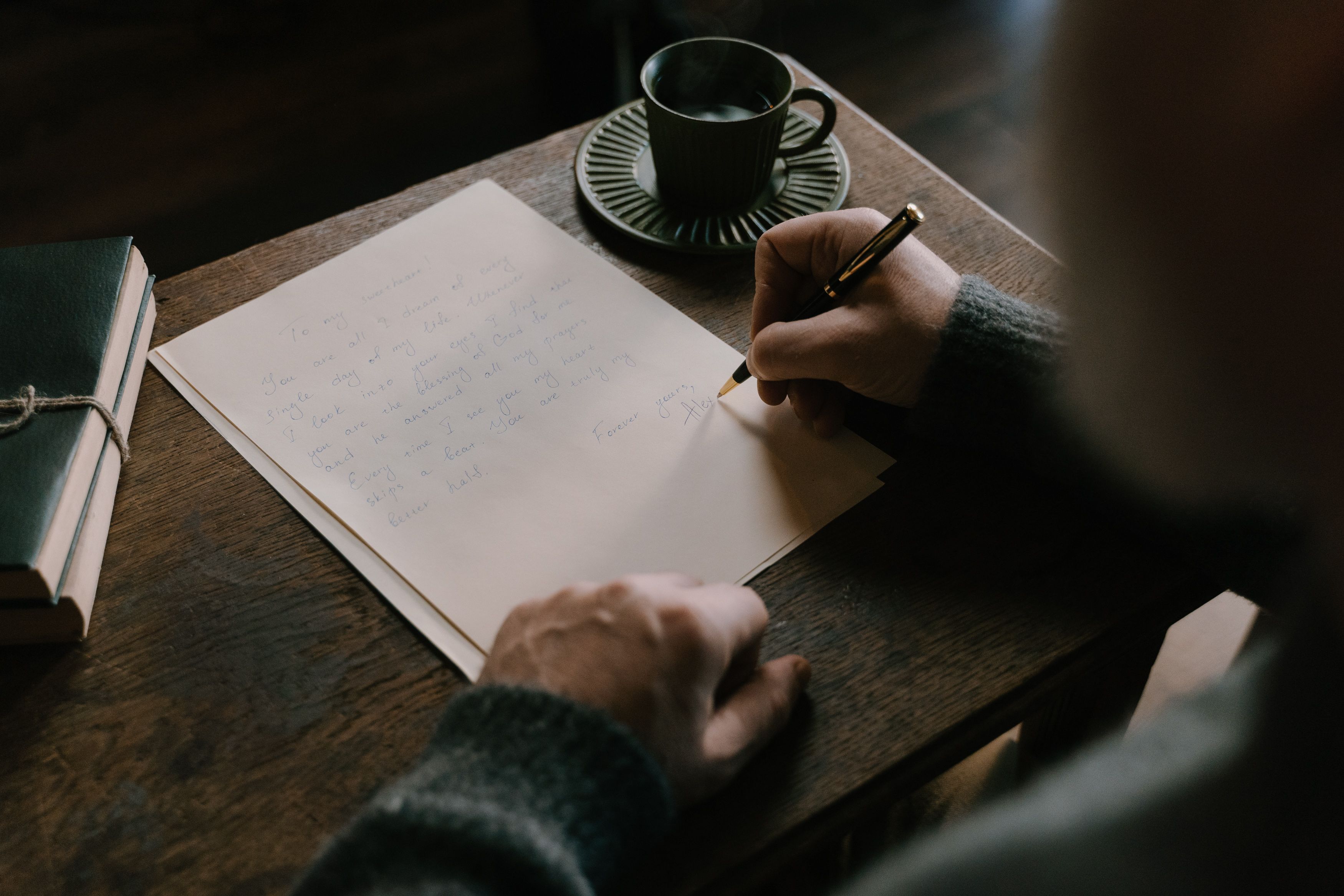 A hand writing a letter with a fountain pen beside a cup of coffee on a wooden desk.